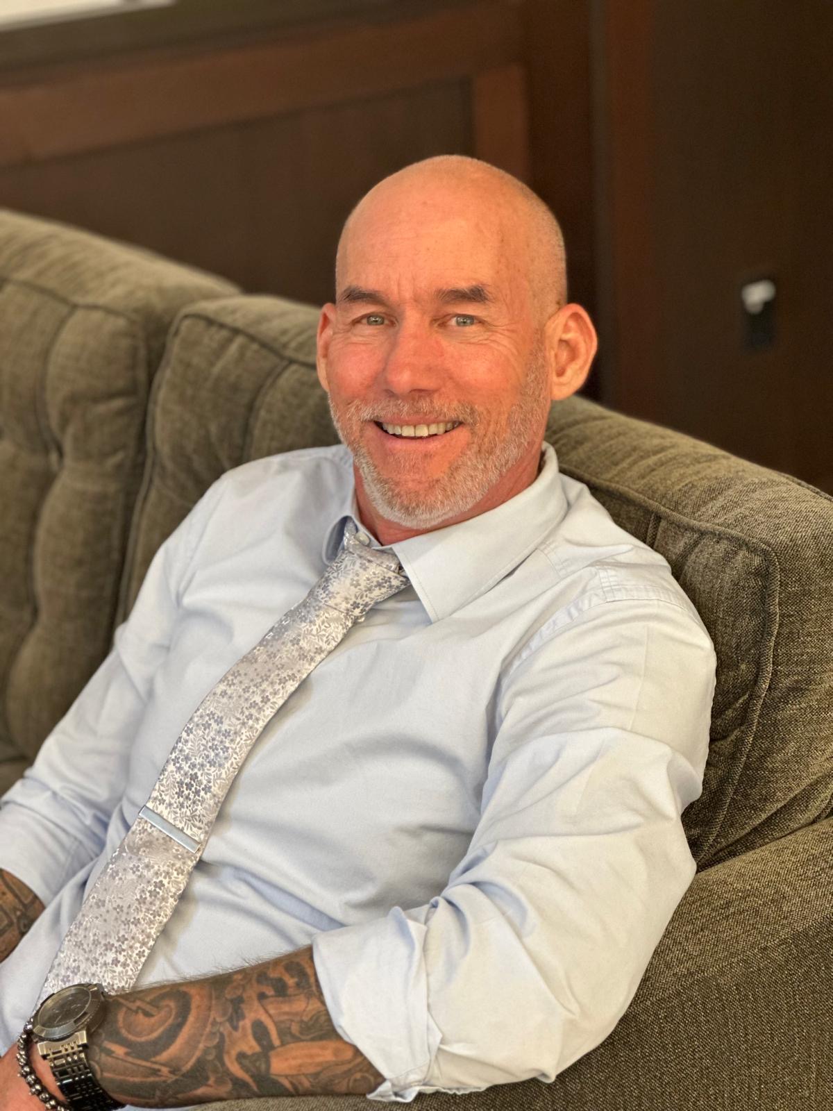 Bald man with a beard smiling, wearing a white shirt and patterned tie, sitting on a sofa.