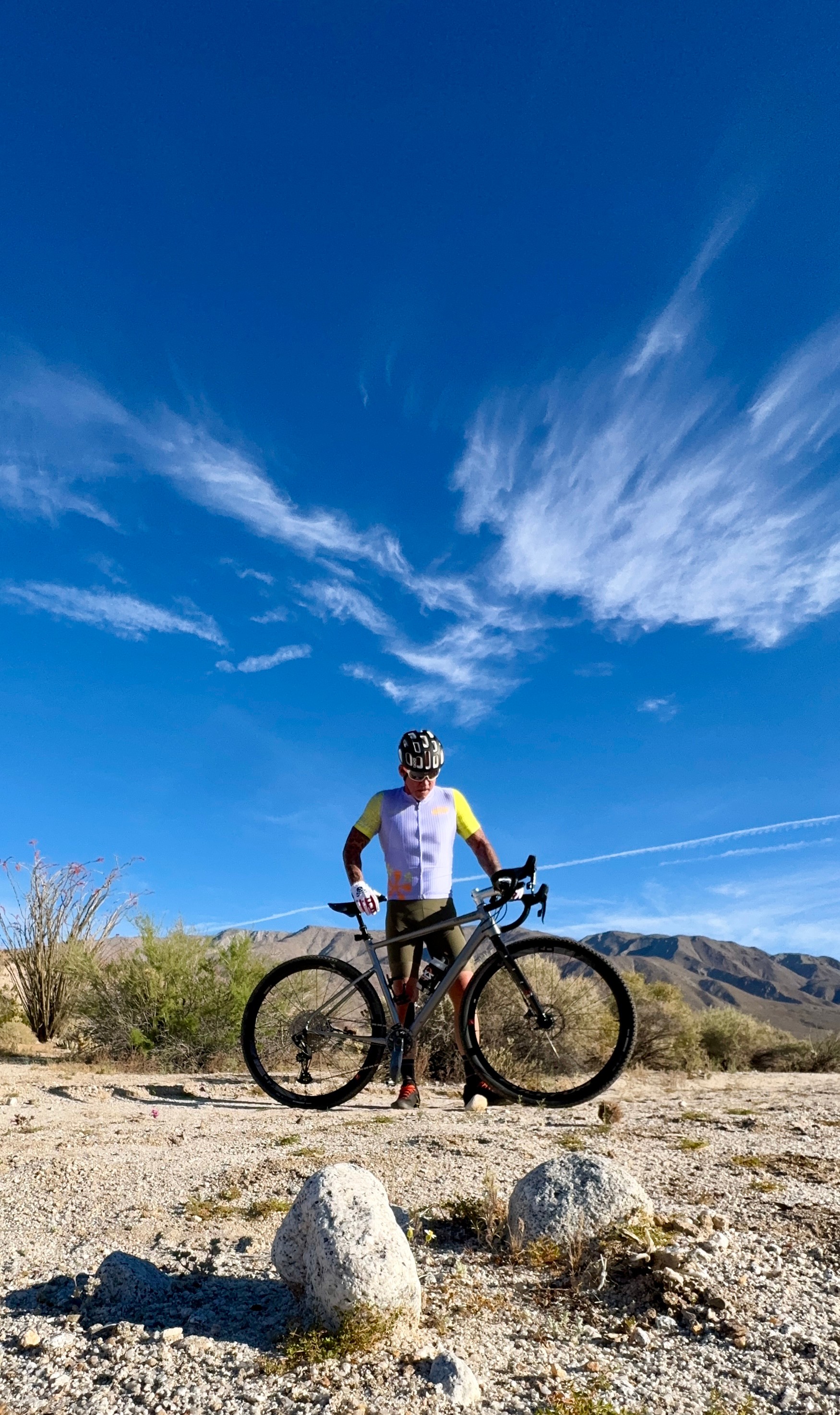 Cyclist standing with bike outdoors under a blue sky with clouds, desert landscape in background, rocks in foreground.