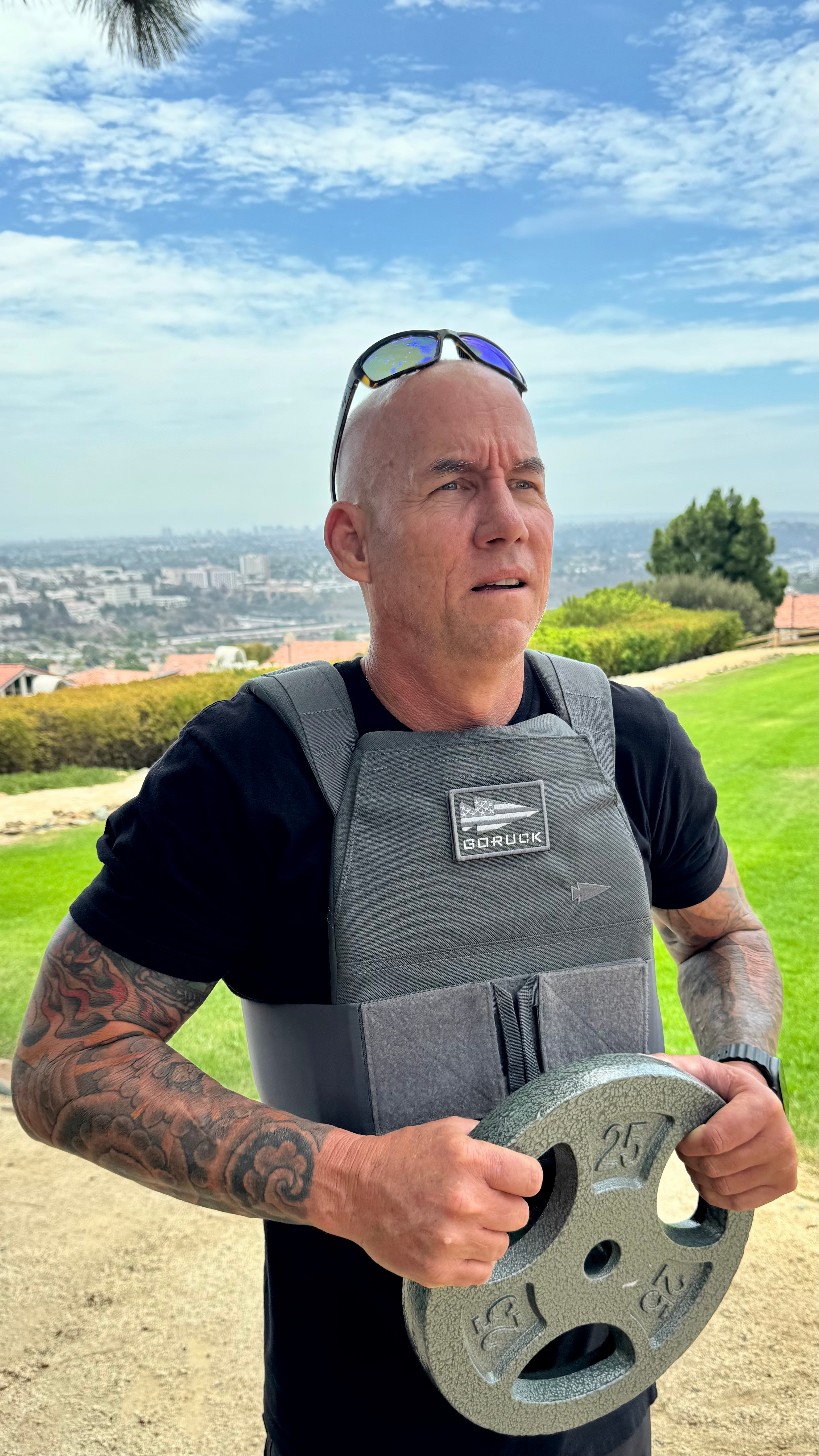Man outdoors holding a circular device, wearing a vest, with a cityscape and blue sky in background.