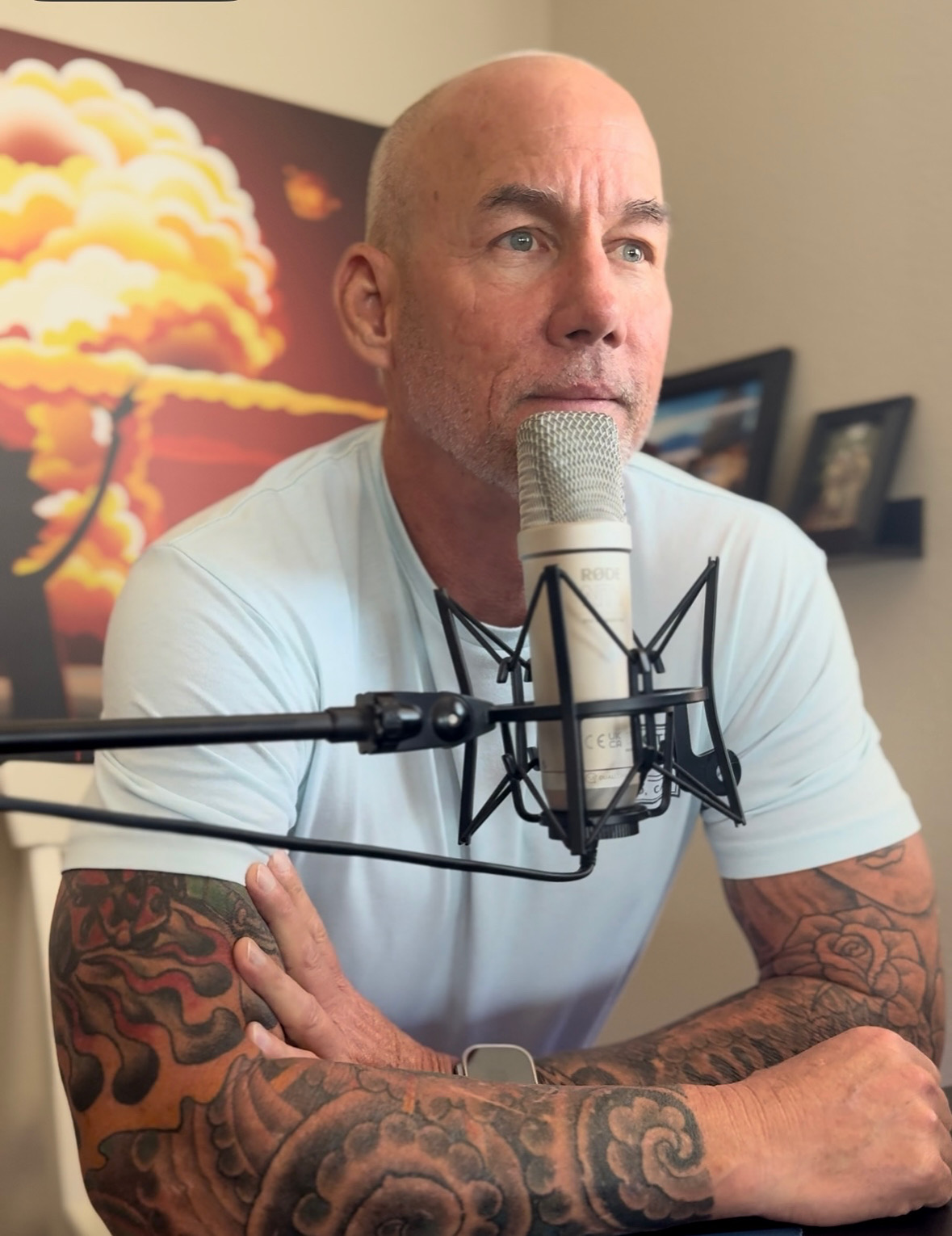 Bald man with tattoos speaking into a microphone, with framed pictures and a colorful cloud painting in the background.