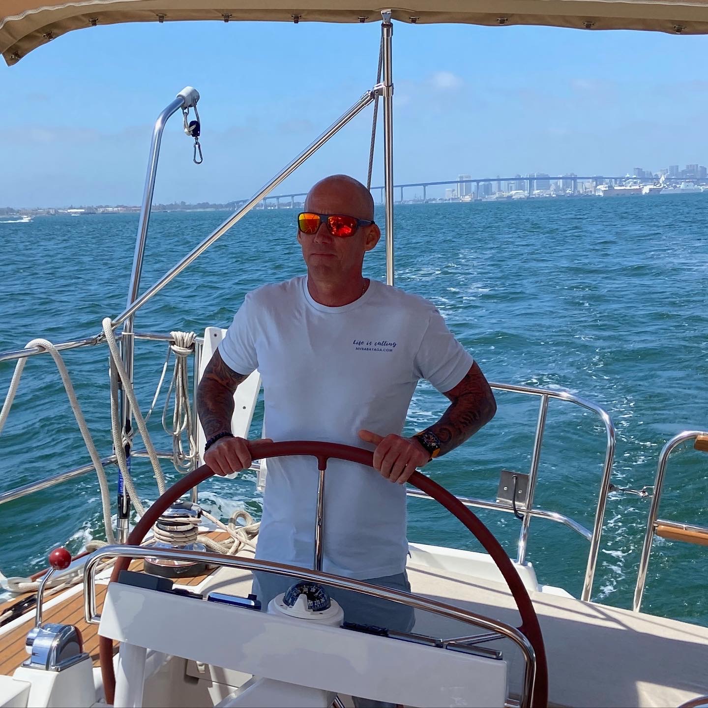 Person standing at the helm of a sailboat holding the steering wheel, wearing sunglasses and a white t-shirt, with water and a city skyline in the background.