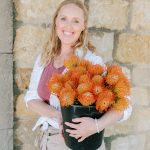 Woman with blonde hair holding a black pot of orange flowers, standing against a stone wall.