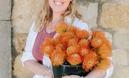 Woman with blonde hair holding a black pot of orange flowers, standing against a stone wall.
