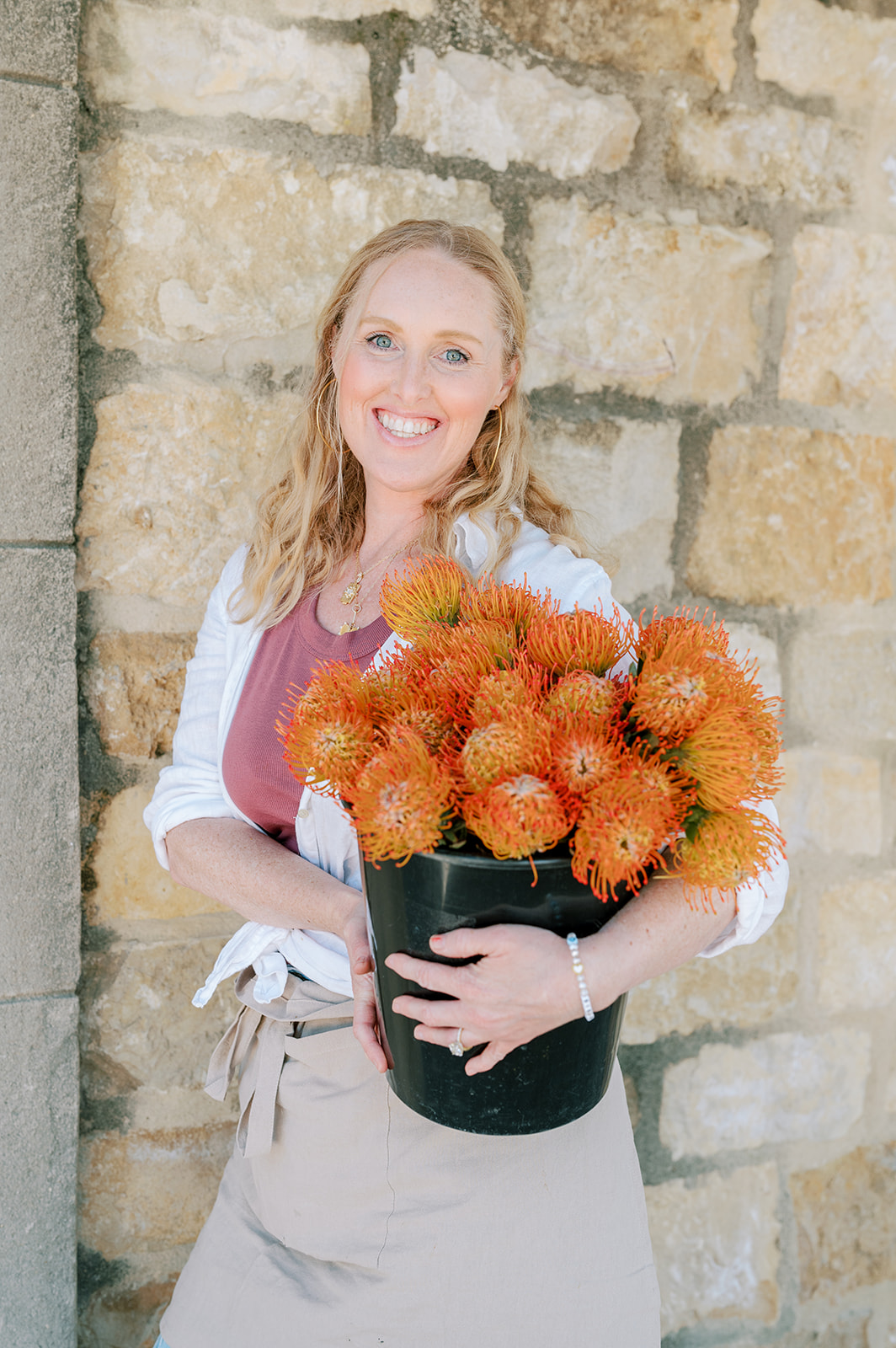 Woman with blonde hair holding a black pot of orange flowers, standing against a stone wall.