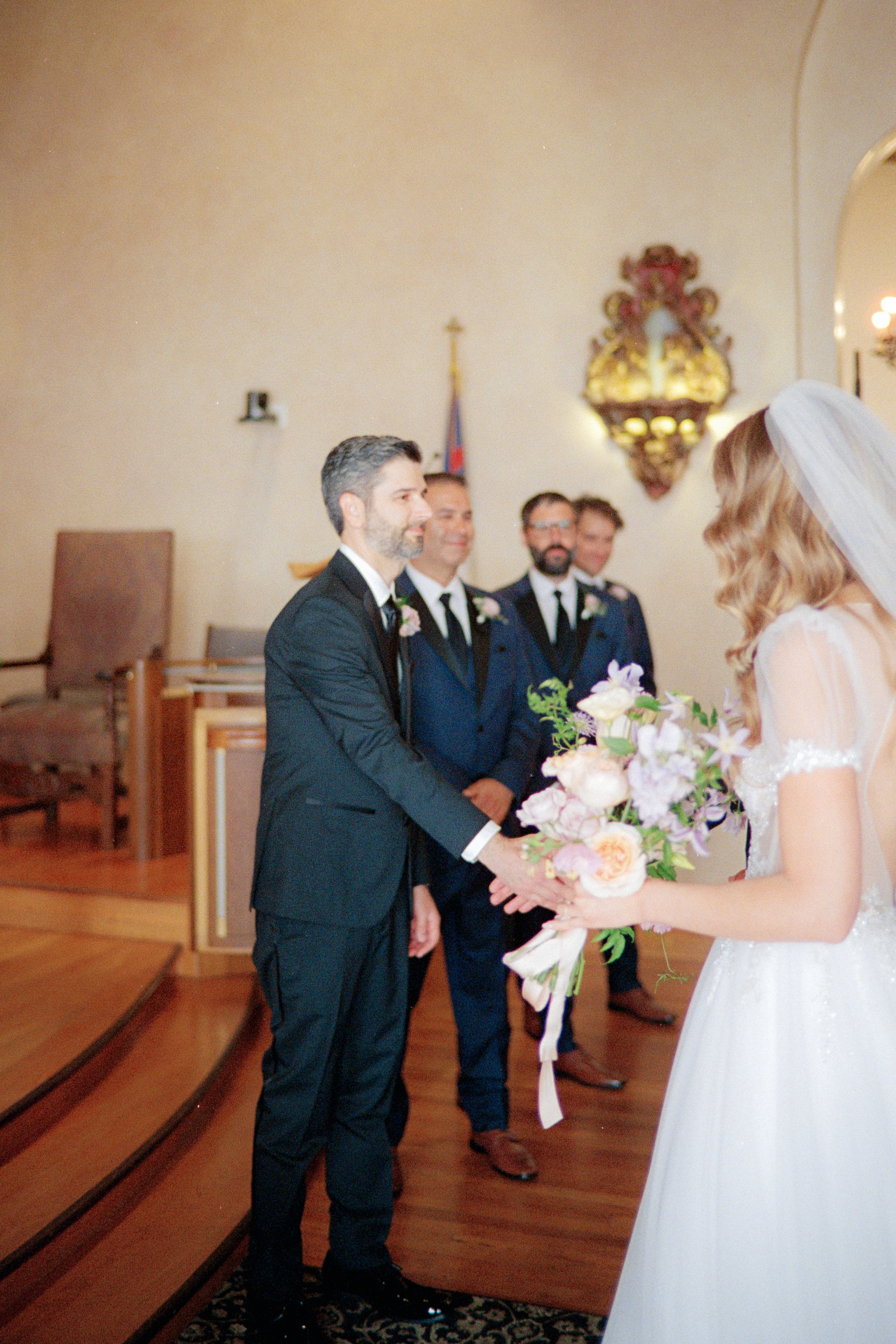 Bride and groom exchanging vows in a church with attendants in the background, holding a bouquet of flowers.