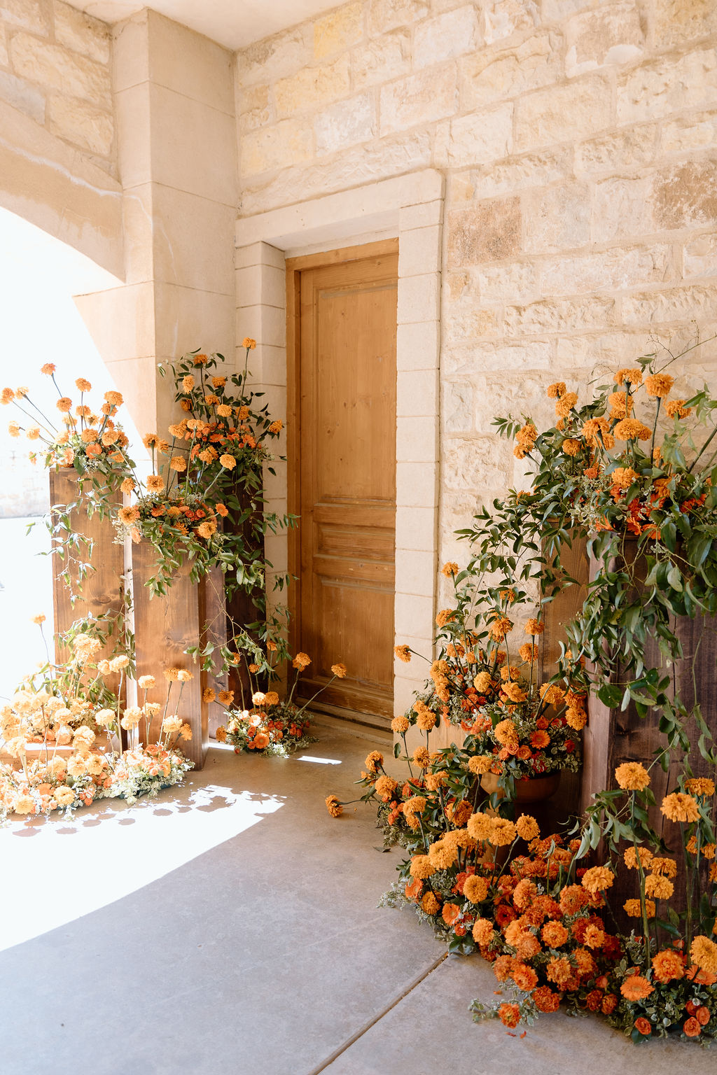 Door surrounded by orange flowers and greenery, with sunlight casting shadows on the stone wall and concrete floor.