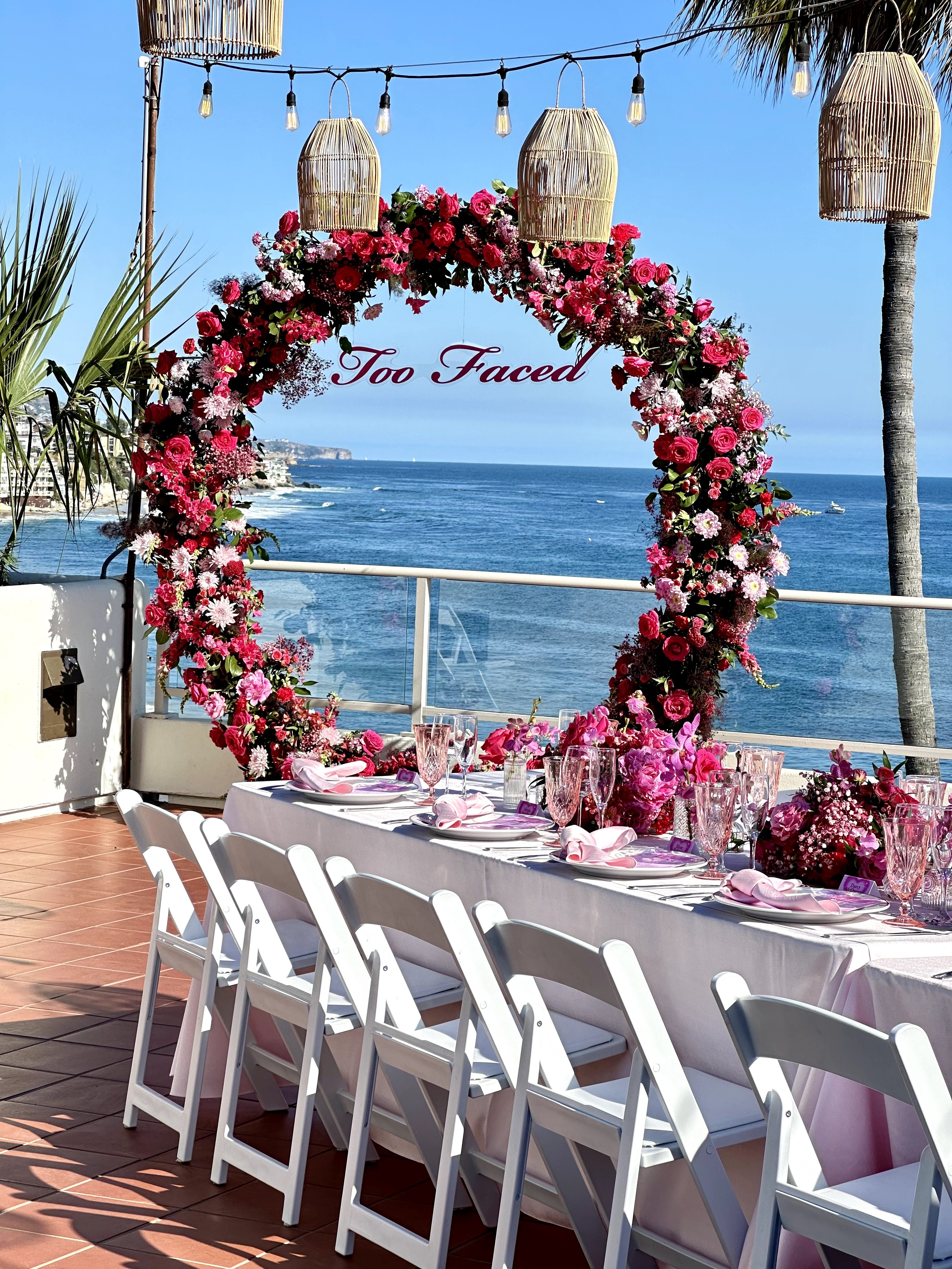 Decorated outdoor dining area with a floral arch, ocean view, and hanging lanterns, set for a celebration or event.