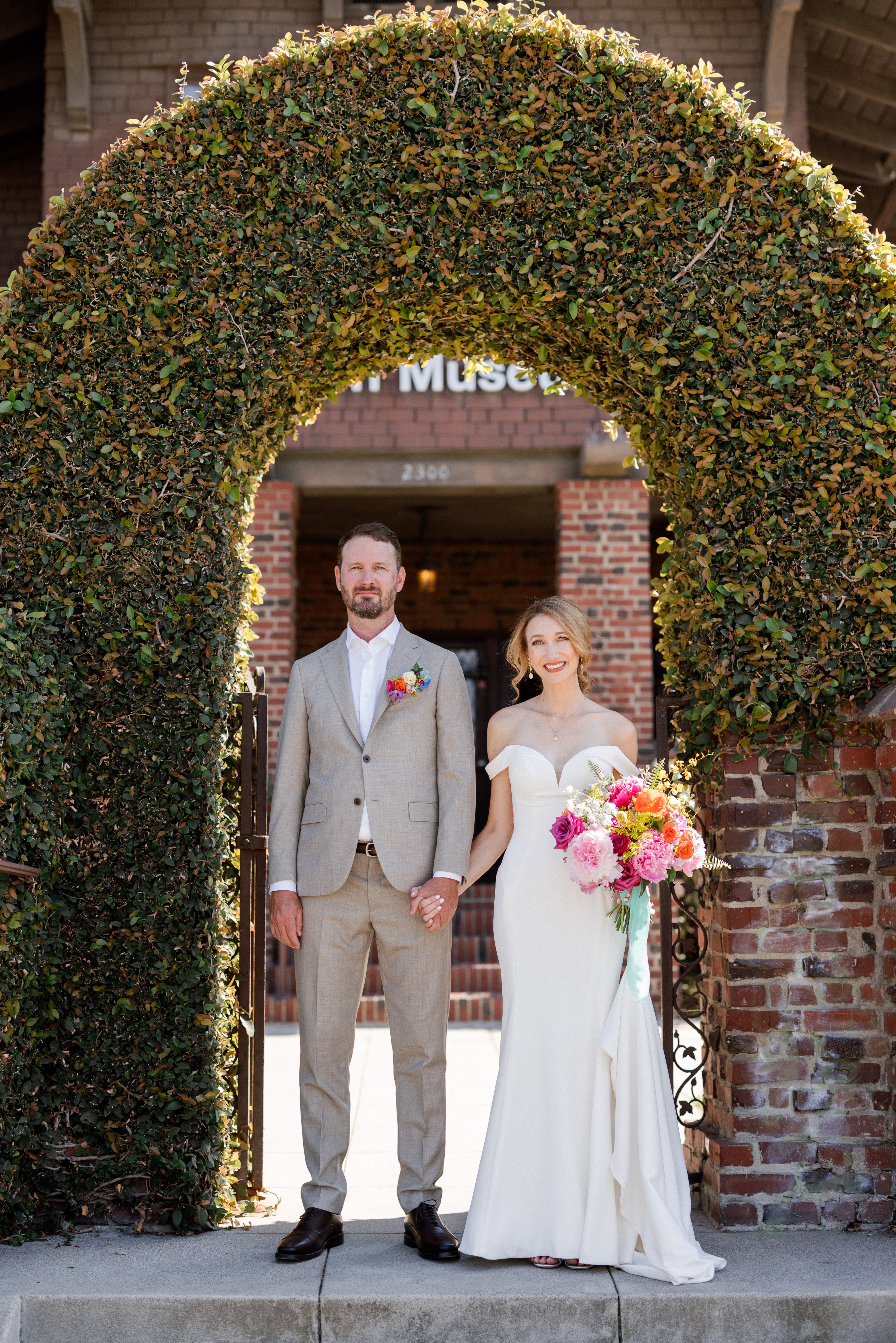A bride and groom stand under a large, green, circular archway made of foliage, holding hands outside a brick building.