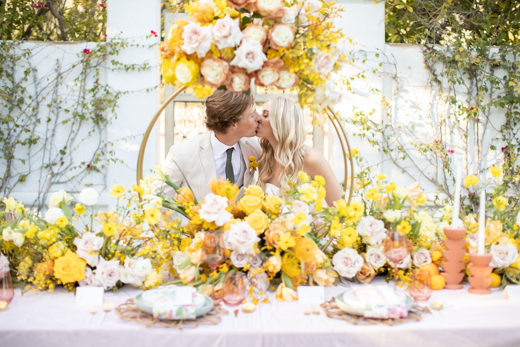 A couple kissing at a decorated table with yellow and white flowers, outdoors with greenery in the background.