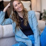 Young woman with long wavy hair smiling, sitting on a sofa in a casual setting, wearing a blue blazer and jeans.