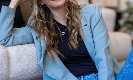 Young woman with long wavy hair smiling, sitting on a sofa in a casual setting, wearing a blue blazer and jeans.