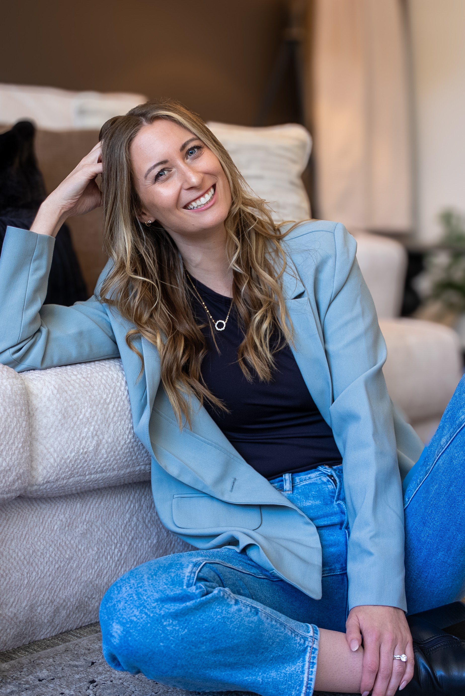 Young woman with long wavy hair smiling, sitting on a sofa in a casual setting, wearing a blue blazer and jeans.