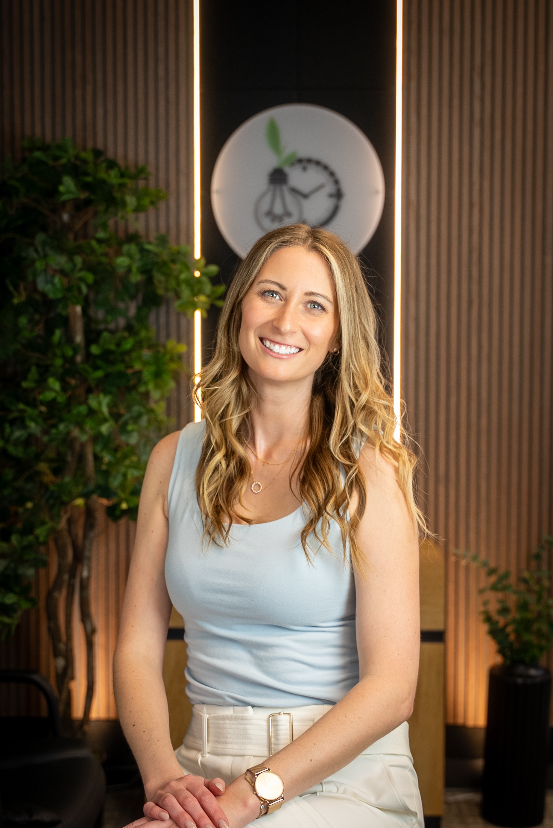 Smiling woman with long wavy hair, wearing a light blue sleeveless top and beige pants, standing indoors with plants and a clock behind her.