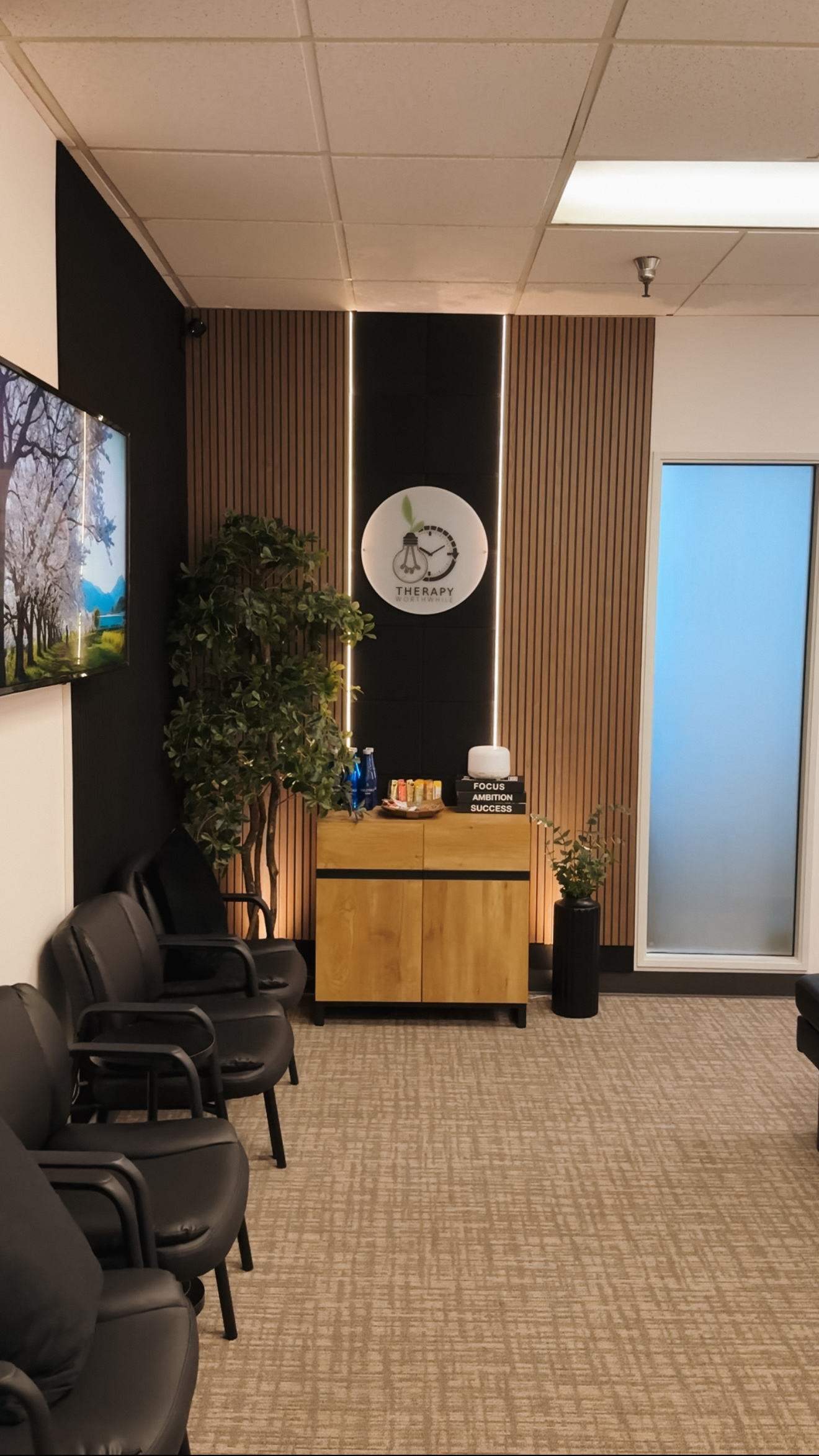Office waiting area with chairs, a wooden cabinet, potted plants, and a wall clock, with a frosted glass door.