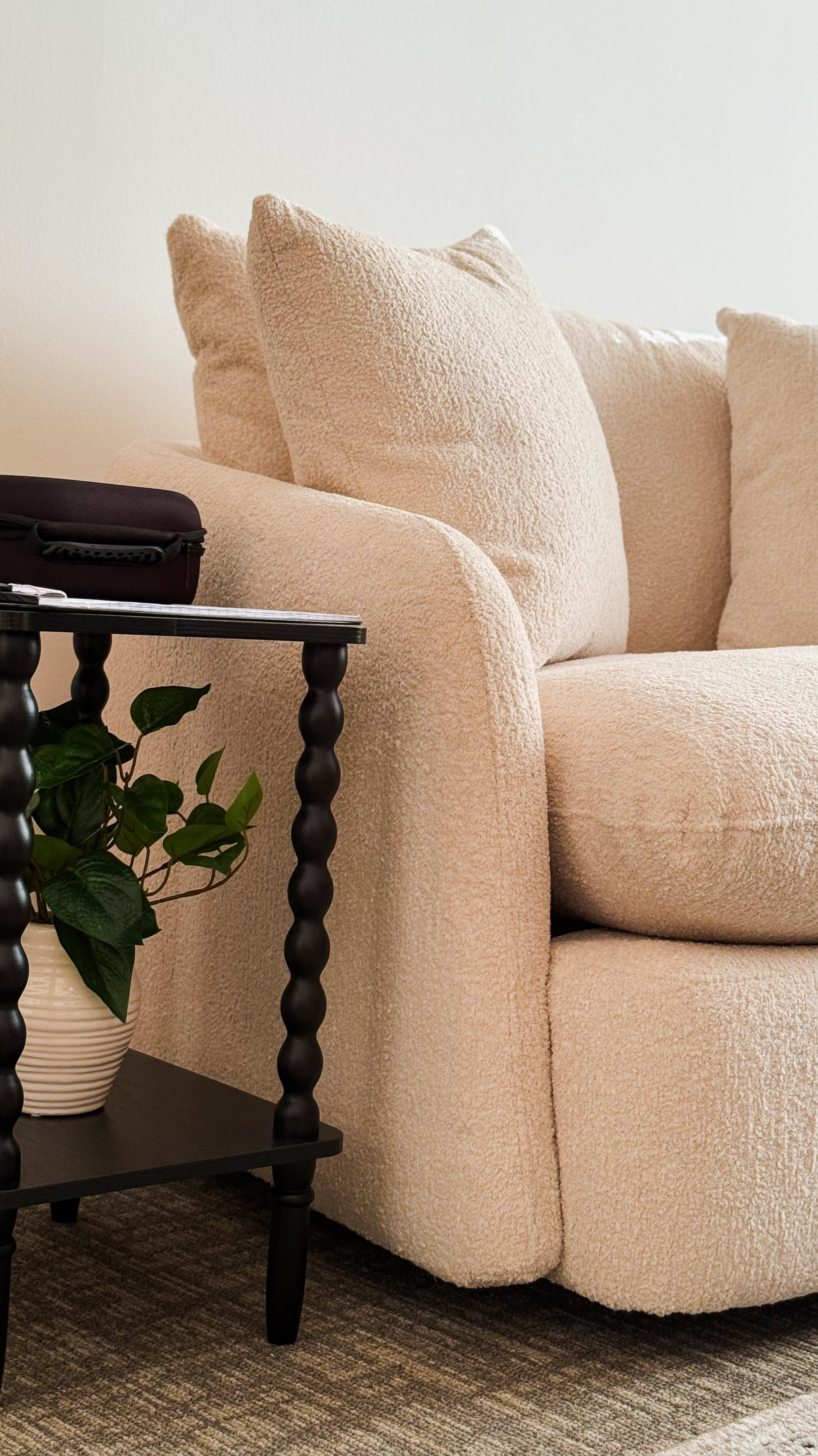 Beige sofa with cushions next to a black side table holding a plant, against a plain wall.