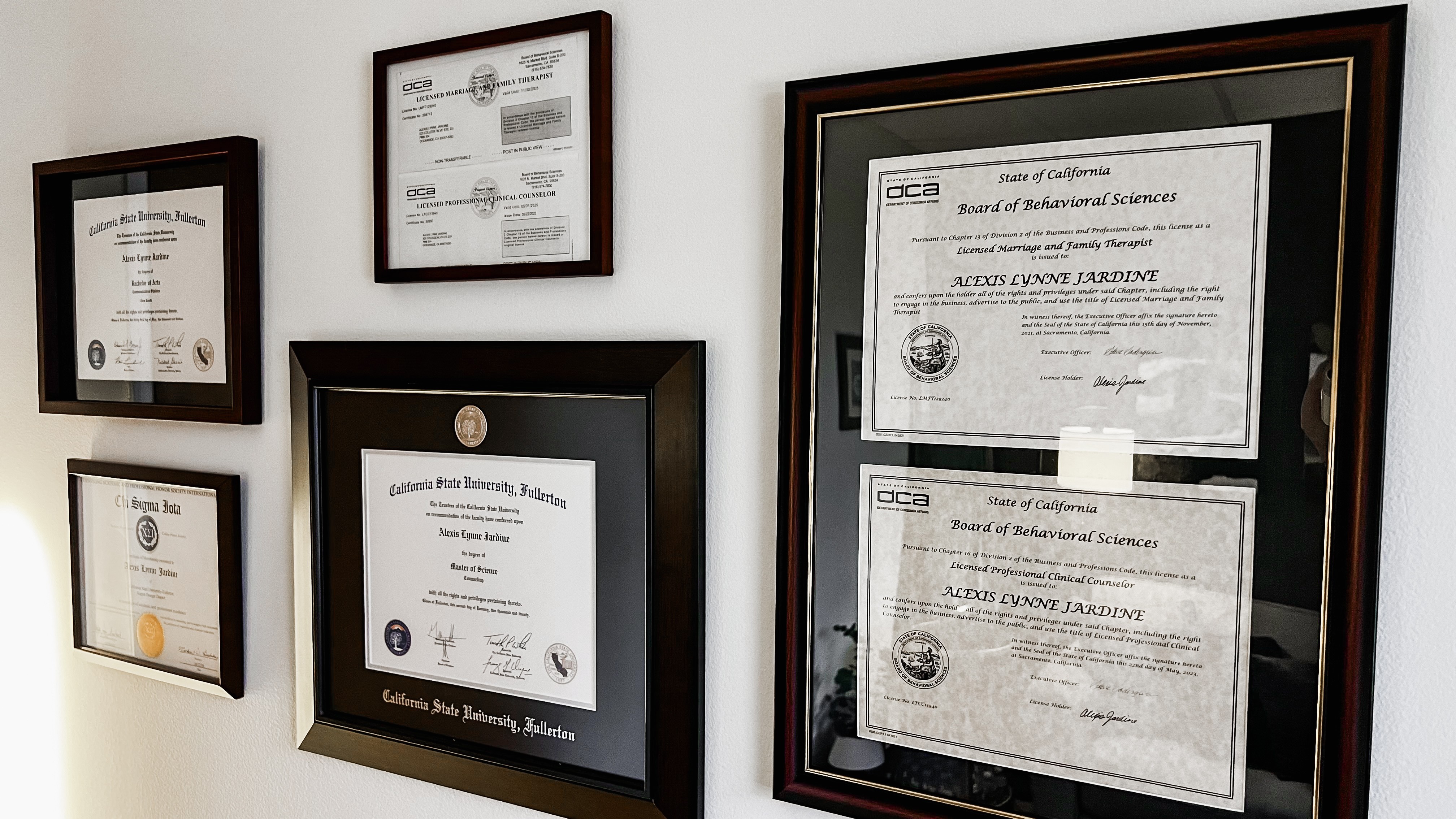 Wall with five framed certificates and diplomas arranged in two rows, some overlapping, on a white wall.